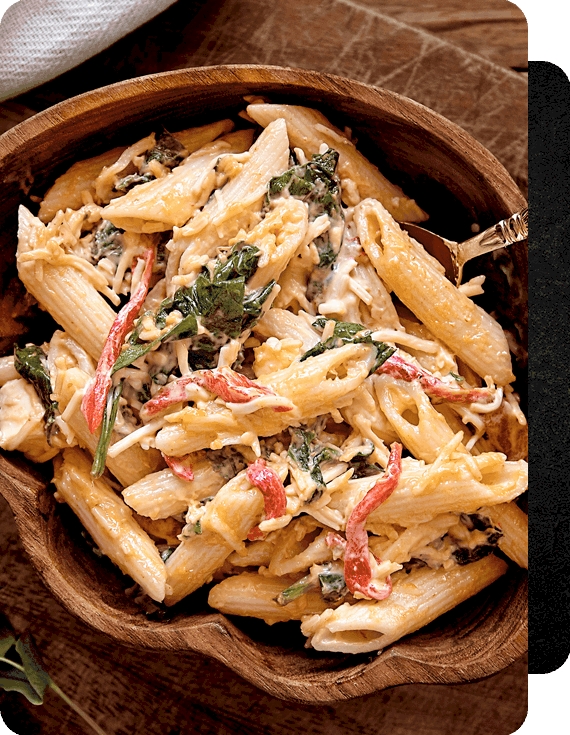 Pasta tipo penne en salsa cremosa, con queso, tiras de pimiento rojo, espinaca, pollo deshebrado, servido en un plato de madera, ideal para tu semana sin estrés. 